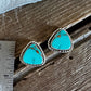 A pair of Southwestern-style turquoise stud earrings with silver bezel settings and twisted rope borders, displayed on a rustic wooden surface next to a metal ruler for scale.