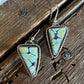 A pair of triangular drop earrings with light blue-green stone inlays and silver-toned bezel settings, shown on a rustic dark wood surface.