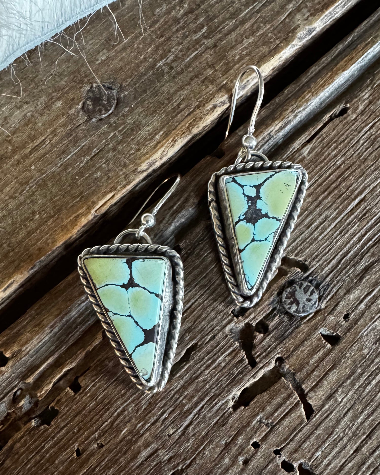A pair of triangular drop earrings with light blue-green stone inlays and silver-toned bezel settings, shown on a rustic dark wood surface.