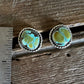 A pair of oval turquoise and gold stud earrings displayed on a rustic wooden surface next to a metal ruler for scale.