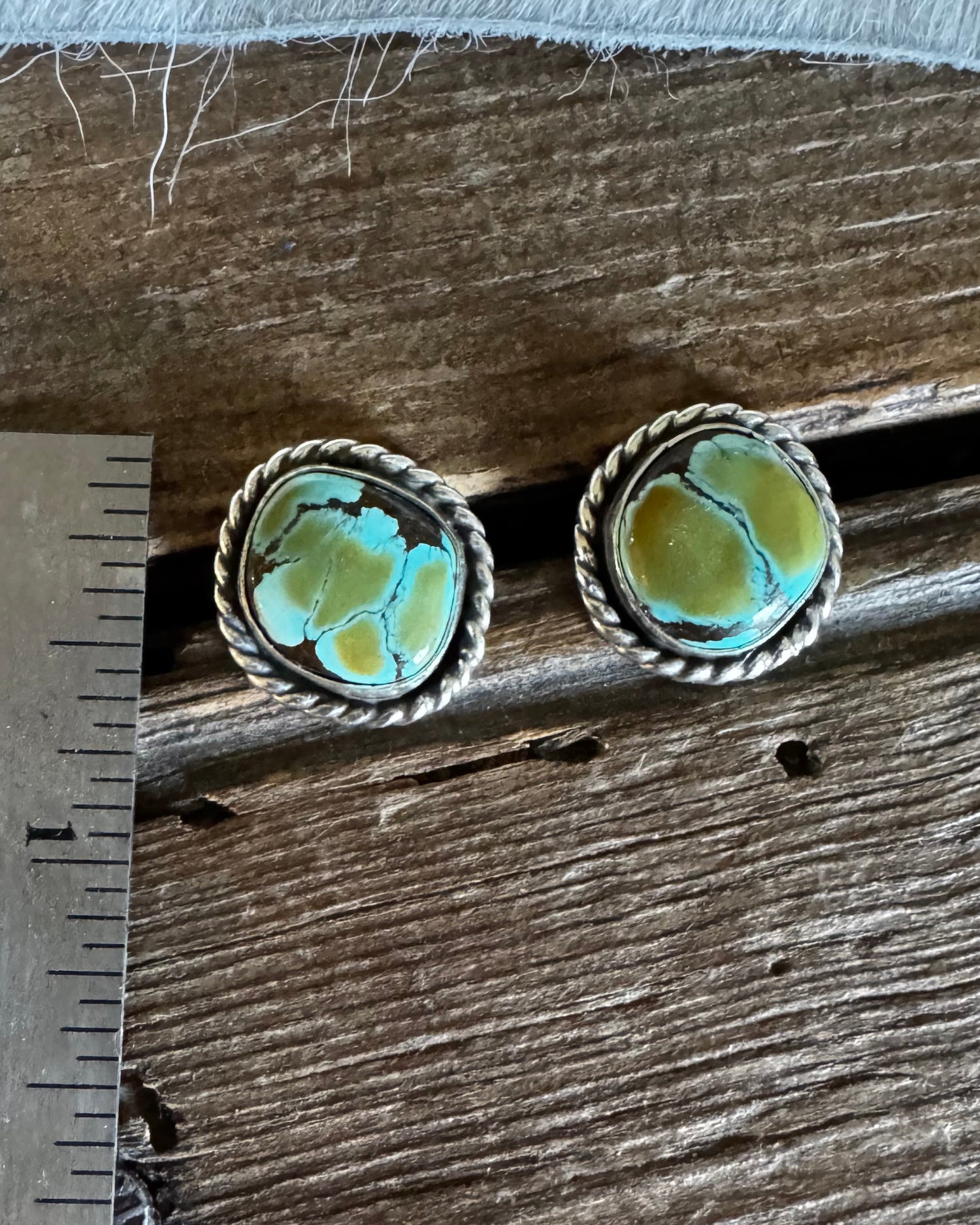 A pair of oval turquoise and gold stud earrings displayed on a rustic wooden surface next to a metal ruler for scale.
