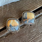 A pair of square stud earrings with silver-toned twisted borders and Maligano Jasper depicting a landscape scene in warm orange and cool blue tones, resting on a rustic wooden surface.
