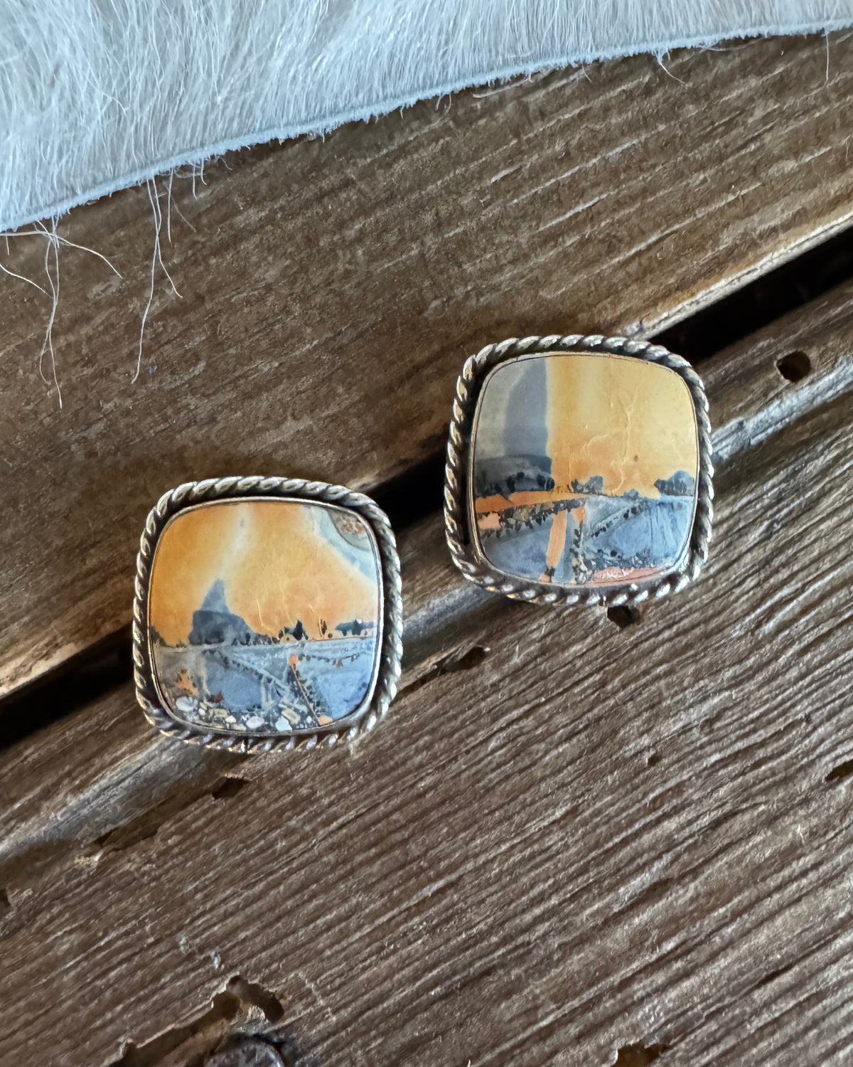 A pair of square stud earrings with silver-toned twisted borders and Maligano Jasper depicting a landscape scene in warm orange and cool blue tones, resting on a rustic wooden surface.