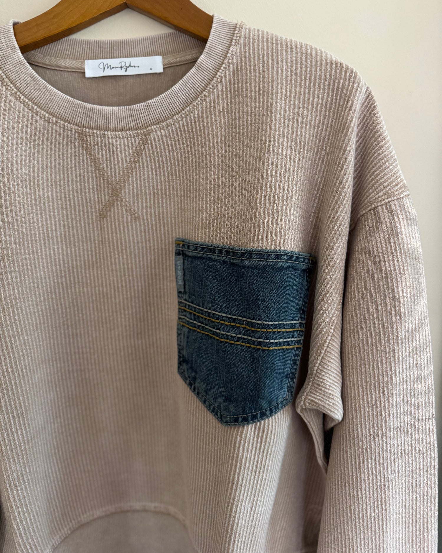 Beige sweatshirt with a denim pocket on a hanger against a beige wall.