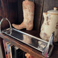 A polished stainless steel serving tray with decorative handles, displayed on a dark wooden shelf next to a cowboy boot and a ceramic teapot.
