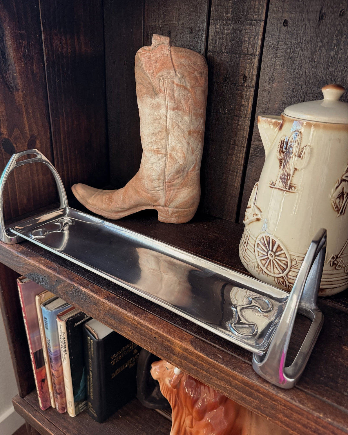 A polished stainless steel serving tray with decorative handles, displayed on a dark wooden shelf next to a cowboy boot and a ceramic teapot.