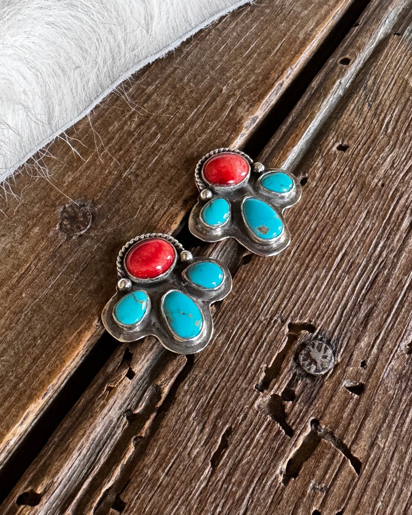 A pair of silver stud earrings with red and turquoise stones, displayed on a rustic wooden surface next to a white faux fur trim.