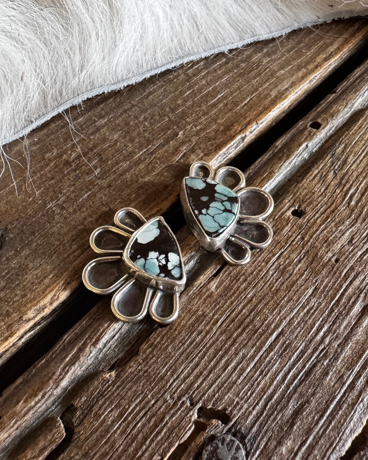 A pair of silver-toned stud earrings with a teardrop-shaped turquoise and black inlay, displayed on a rustic wooden surface next to white faux fur.