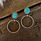 A pair of bohemian-style dangle earrings with oval turquoise-blue stones in gold settings and large twisted hoop drops, displayed on a rustic dark wood surface with a white faux fur accent.