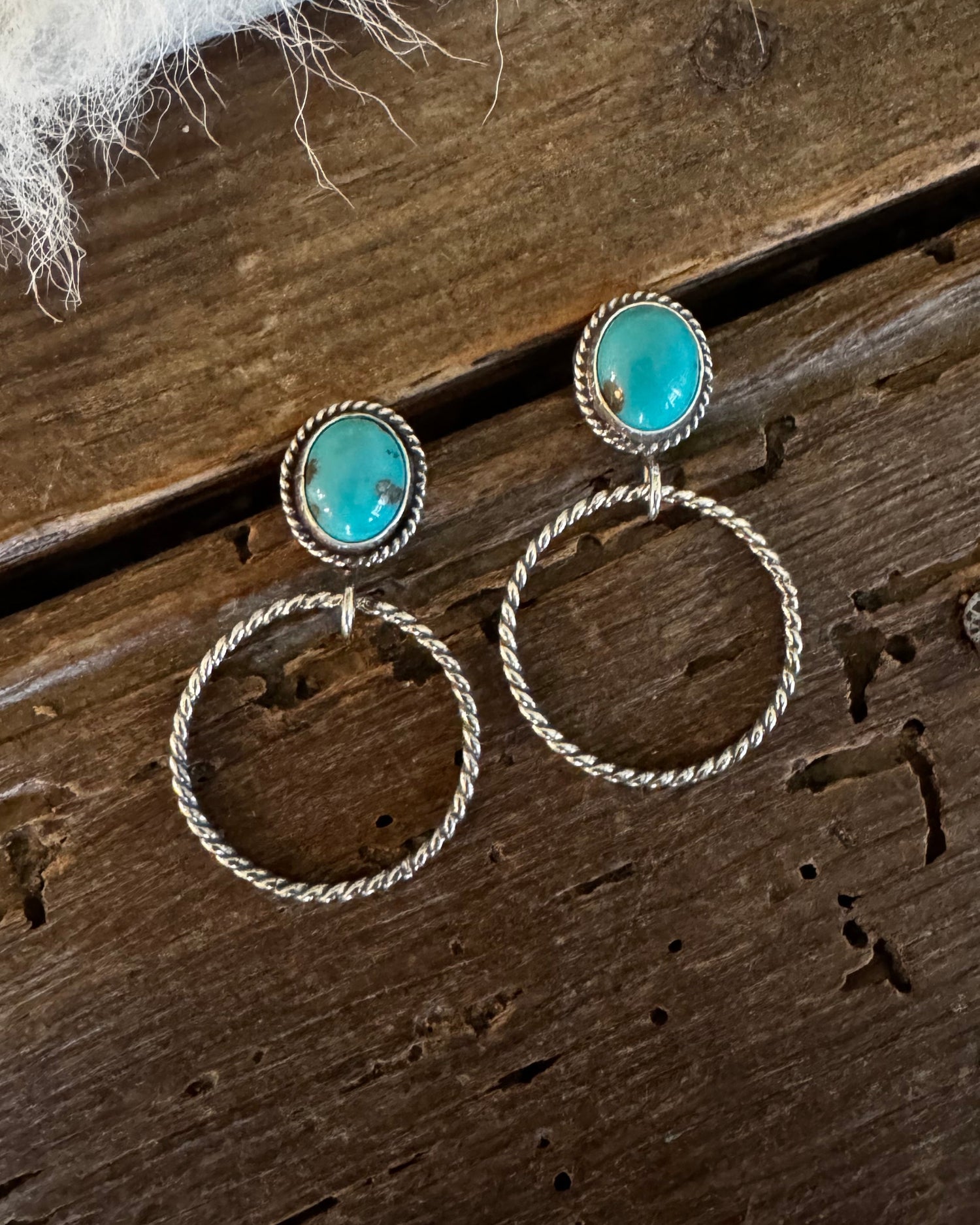 A pair of bohemian-style dangle earrings with oval turquoise-blue stones in gold settings and large twisted hoop drops, displayed on a rustic dark wood surface with a white faux fur accent.