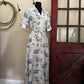 White dress with blue patterns on a mannequin against a rustic background with a red door and wooden wall.