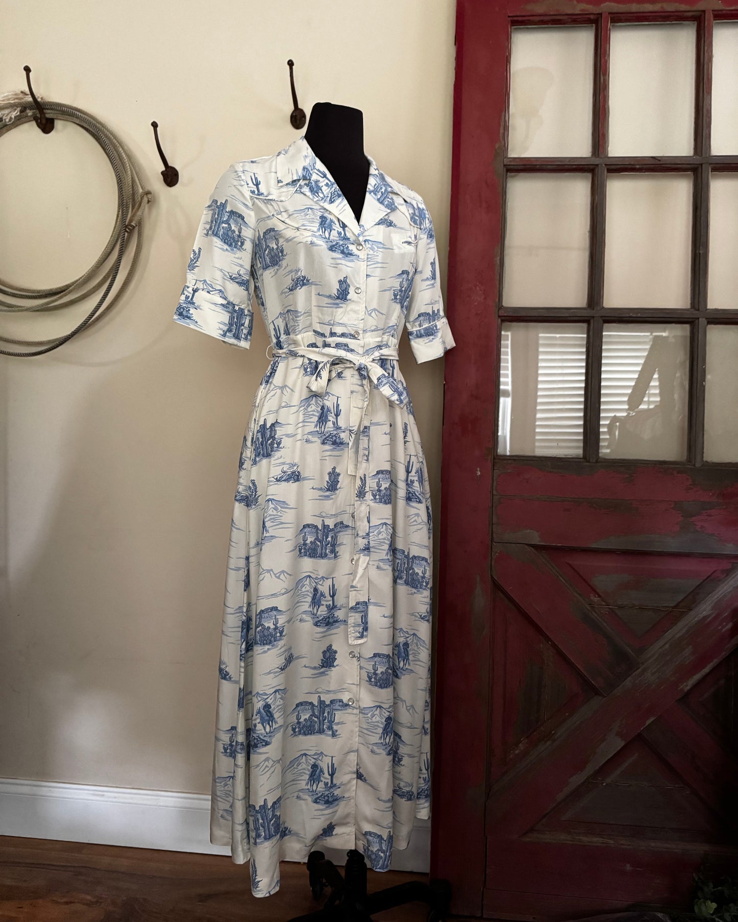 White dress with blue patterns on a mannequin against a rustic background with a red door and wooden wall.