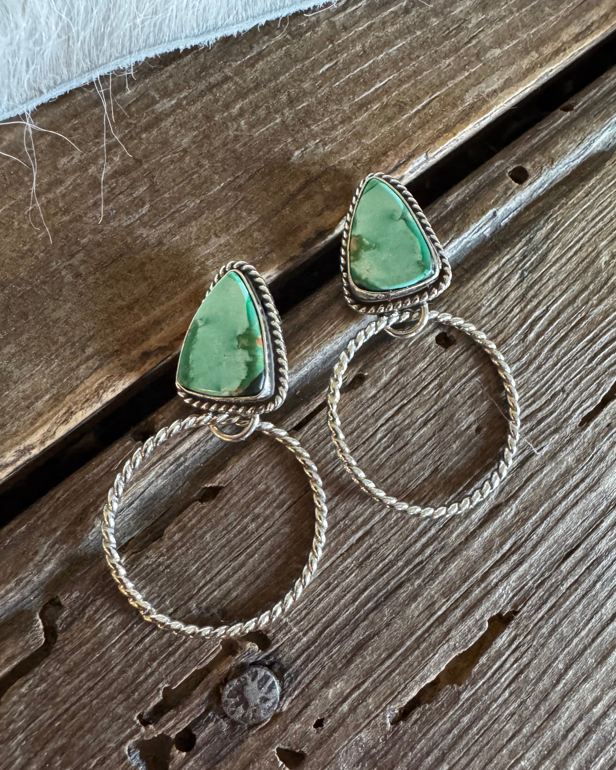 A pair of handmade silver dangle earrings with triangular green stone tops and large twisted hoop drops, displayed on a rustic wooden surface.