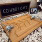A rectangular bamboo serving board with a carved geometric design and two silver metal cowboy handle grips, displayed on a granite countertop in front of a chalkboard sign that reads 'COWBOY CAFE'.