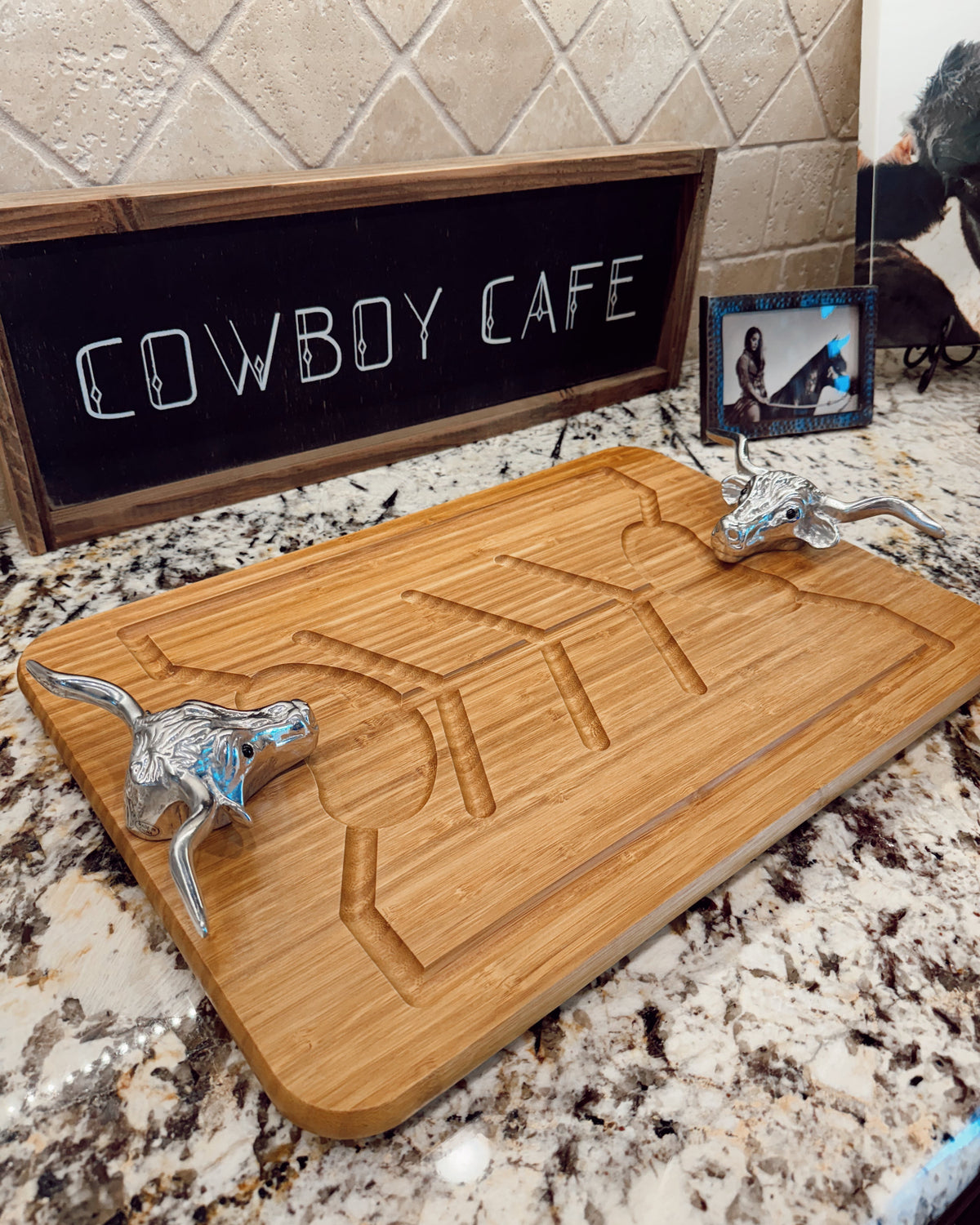 A rectangular bamboo serving board with a carved geometric design and two silver metal cowboy handle grips, displayed on a granite countertop in front of a chalkboard sign that reads 'COWBOY CAFE'.