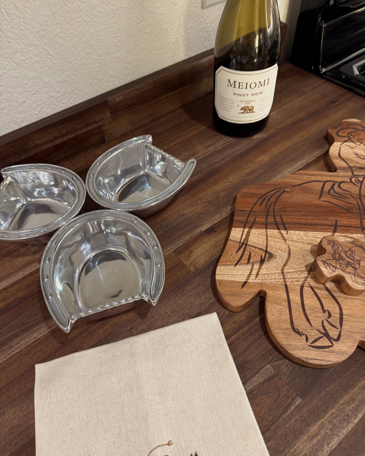 Three silver wine coasters on a wooden surface with a bottle of Meiomi wine and a wooden cutting board.