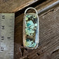 A rectangular turquoise stone pendant with a silver-toned bezel and loop, shown next to a metal ruler on a rustic wooden surface.