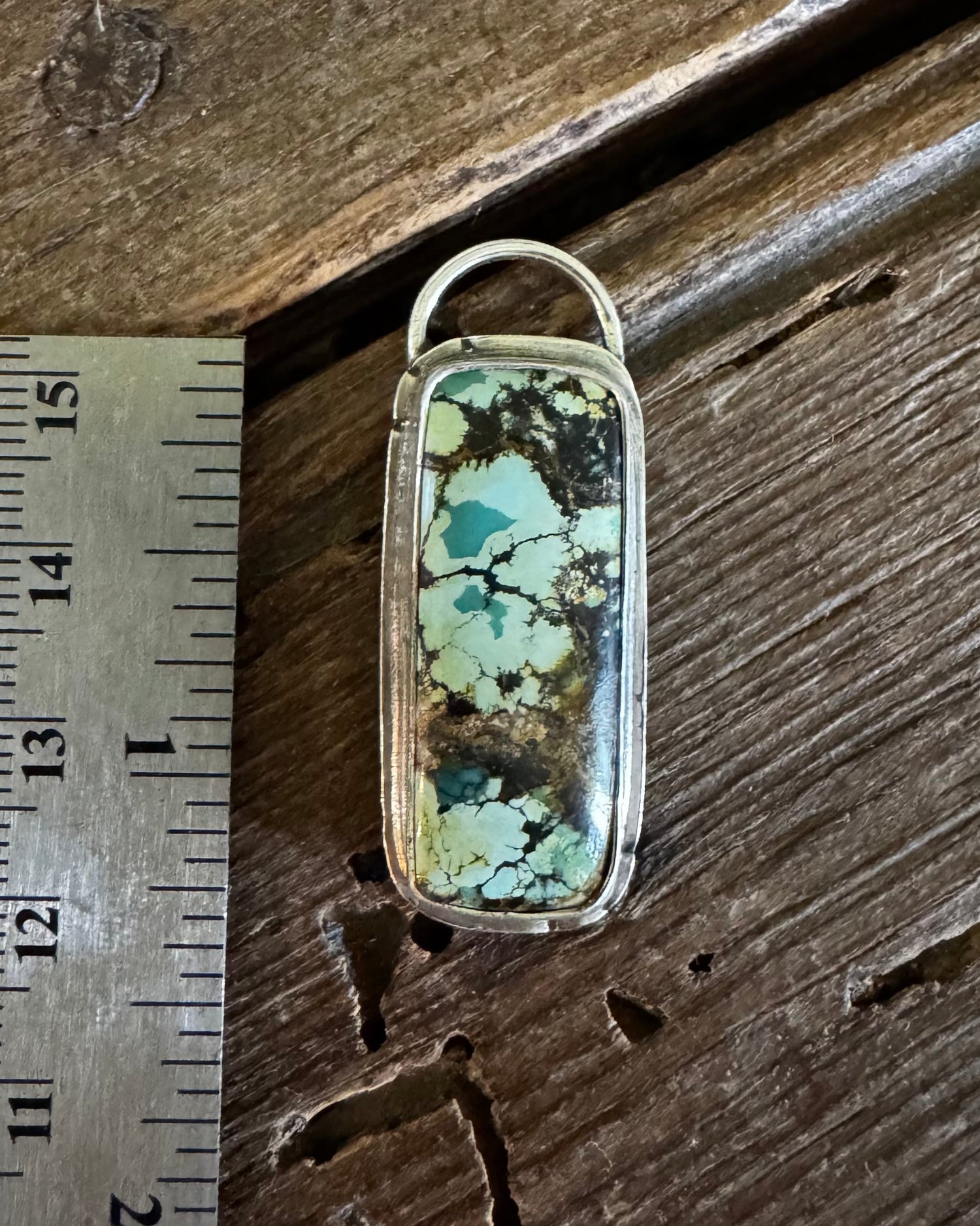 A rectangular turquoise stone pendant with a silver-toned bezel and loop, shown next to a metal ruler on a rustic wooden surface.