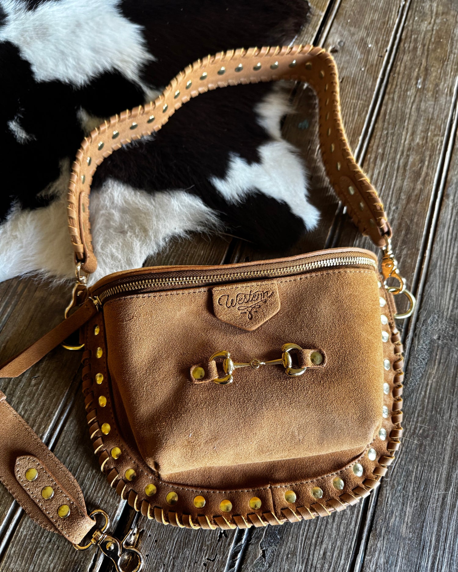 Brown leather crossbody bag with gold accents on a wooden surface with cowhide rug