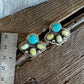 A pair of silver paw-shaped stud earrings with turquoise and yellow-green stone accents, displayed on a rustic wooden surface next to a metal ruler for scale.