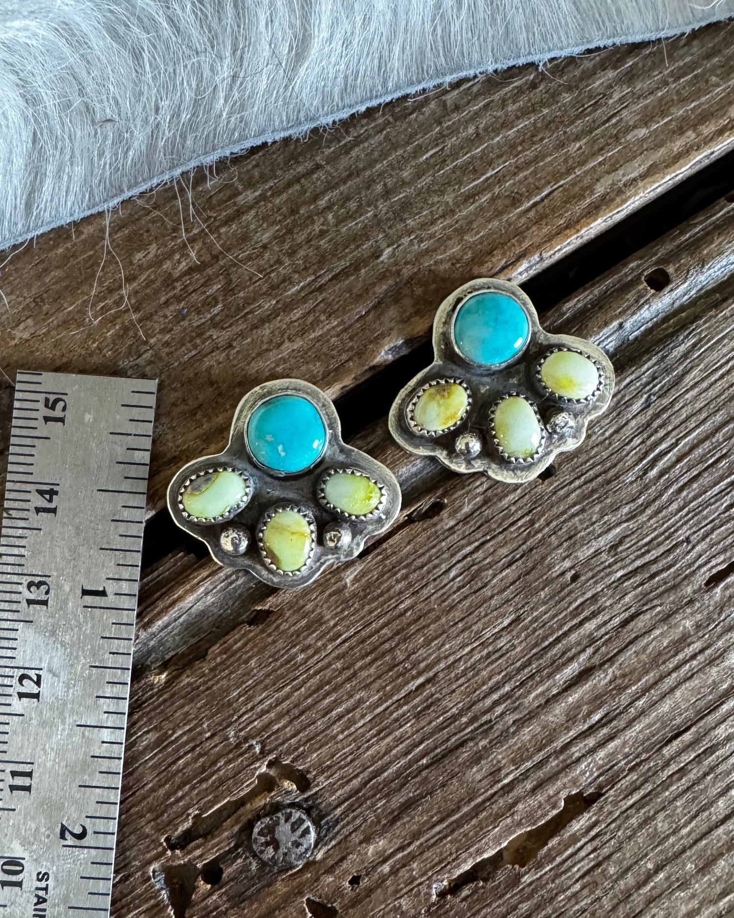A pair of silver paw-shaped stud earrings with turquoise and yellow-green stone accents, displayed on a rustic wooden surface next to a metal ruler for scale.