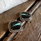 A pair of silver-toned stud earrings with rectangular black stone inlays featuring a bright turquoise-blue stripe, displayed on a rustic wooden surface with a white faux-fur trim in the background.