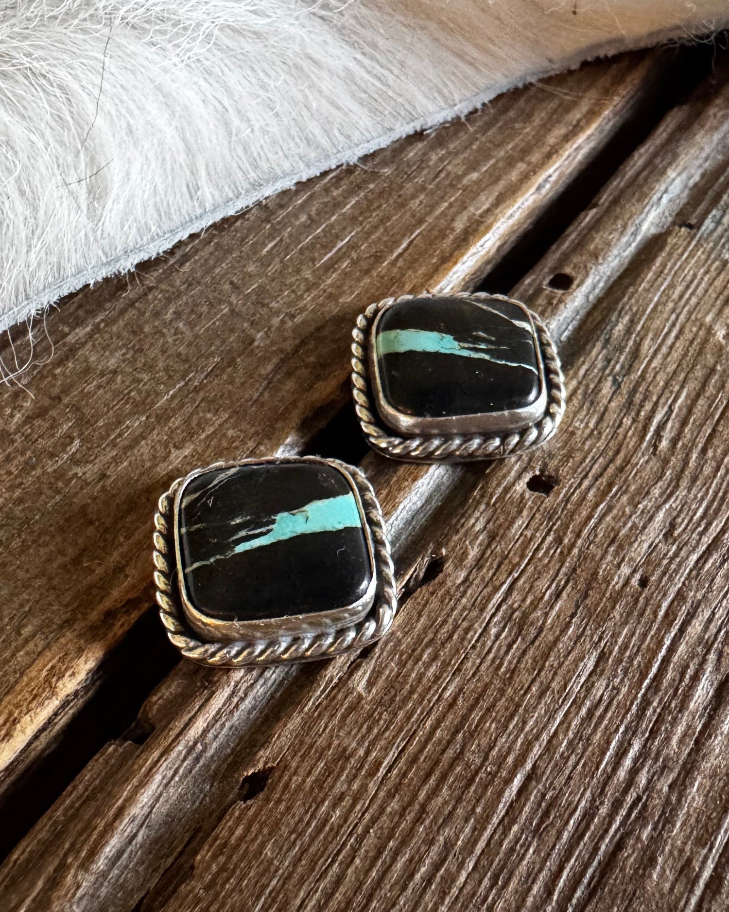 A pair of silver-toned stud earrings with rectangular black stone inlays featuring a bright turquoise-blue stripe, displayed on a rustic wooden surface with a white faux-fur trim in the background.