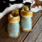 A pair of slip-on slippers with turquoise floral embossed uppers, tan soles, and fluffy tan faux-fur trim, displayed on a rustic wooden surface with a black-and-white cowhide rug in the background.