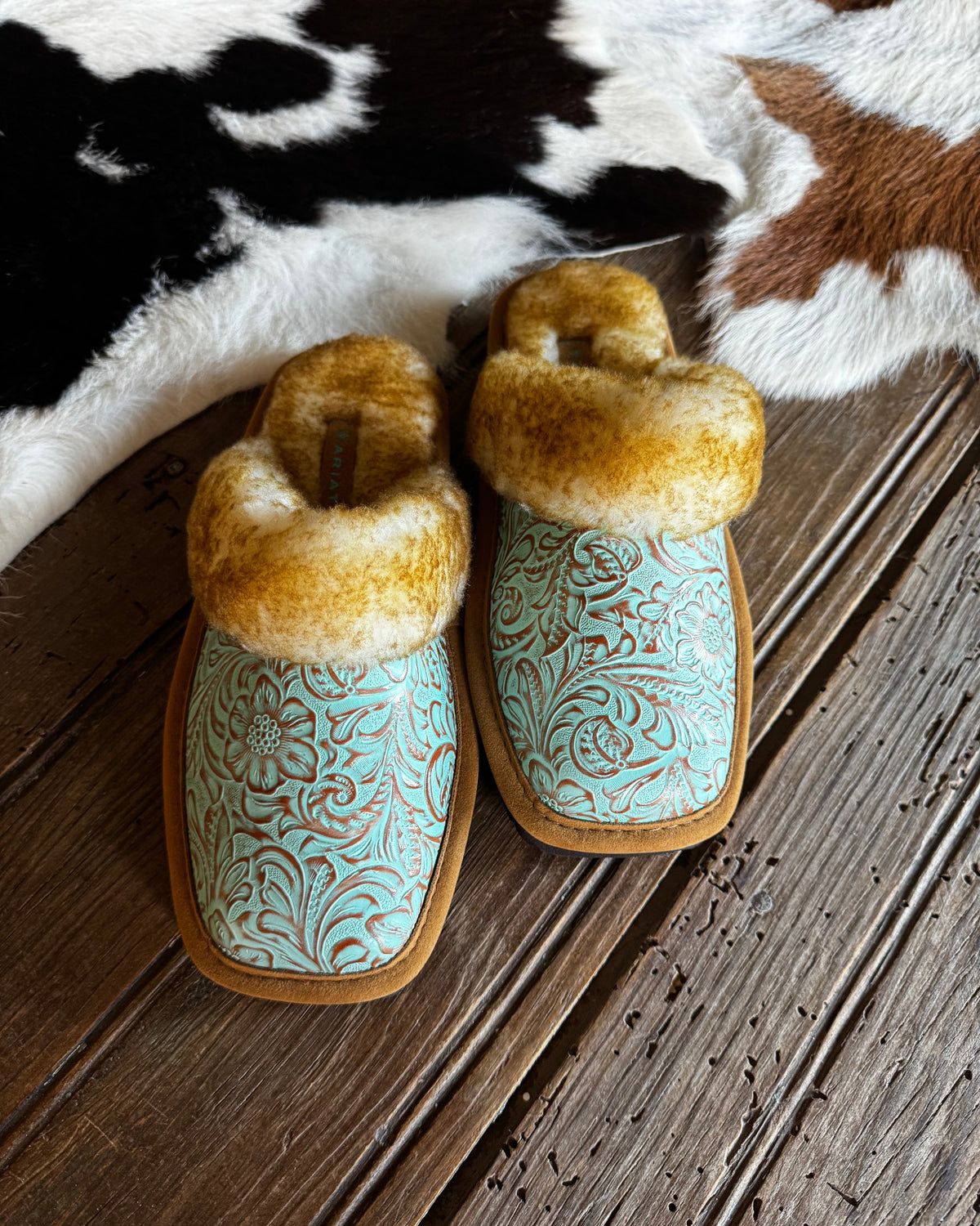 A pair of slip-on slippers with turquoise floral embossed uppers, tan soles, and fluffy tan faux-fur trim, displayed on a rustic wooden surface with a black-and-white cowhide rug in the background.