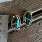 A pair of silver-toned stud earrings with oval black stones featuring bright turquoise blue matrix, displayed on a rustic wooden surface next to a metal ruler for scale.