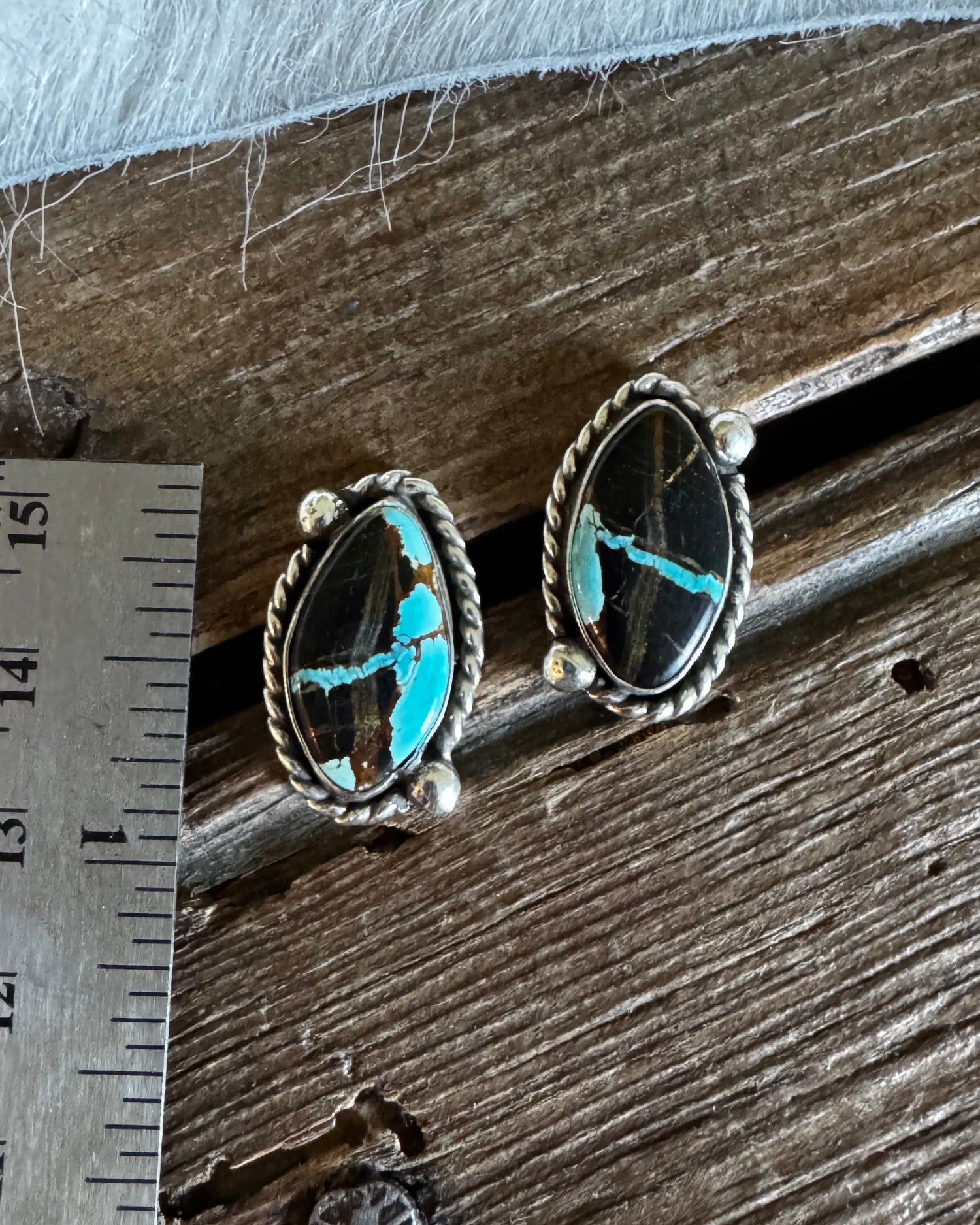 A pair of silver-toned stud earrings with oval black stones featuring bright turquoise blue matrix, displayed on a rustic wooden surface next to a metal ruler for scale.