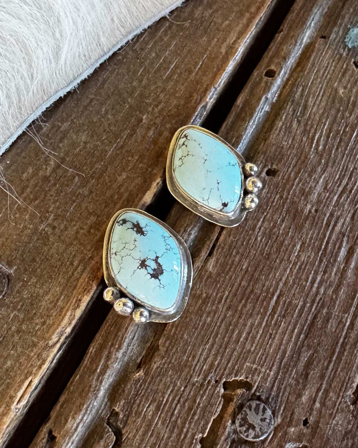 A pair of turquoise statement earrings with gold settings, displayed on a rustic wooden surface next to a white fur trim.