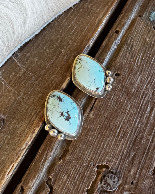 A pair of turquoise statement earrings with gold settings, displayed on a rustic wooden surface next to a white fur trim.