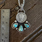 A silver pendant necklace with a coin charm and turquoise stones, displayed next to a metal ruler on a dark wooden surface.