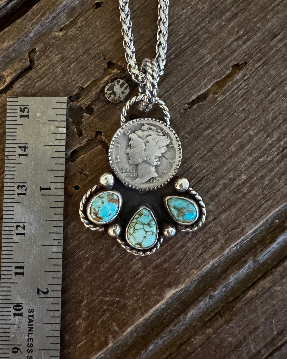 A silver pendant necklace with a coin charm and turquoise stones, displayed next to a metal ruler on a dark wooden surface.