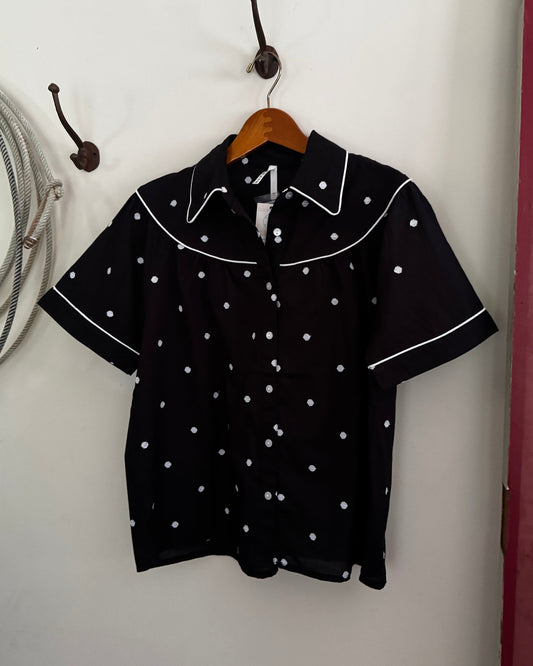 Black blouse with white polka dots on a hanger against a white wall.