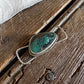 A silver-toned ring with a teardrop-shaped turquoise stone, displayed on a rustic wooden surface with a white faux fur trim at the top.