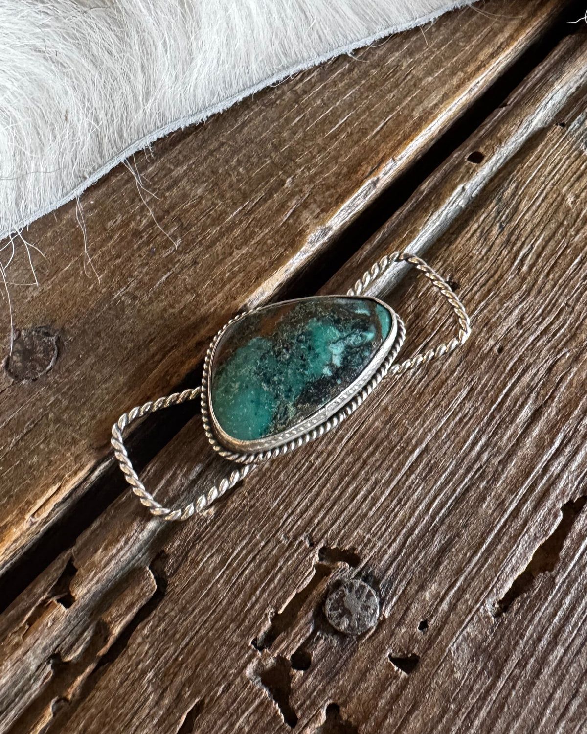 A silver-toned ring with a teardrop-shaped turquoise stone, displayed on a rustic wooden surface with a white faux fur trim at the top.