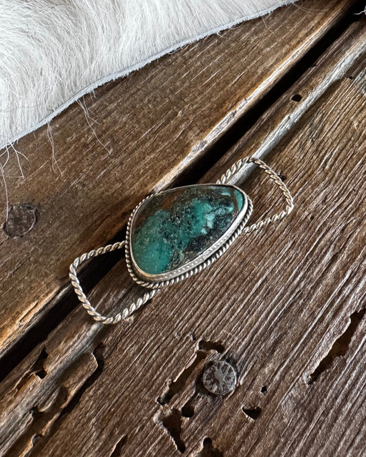 A silver-toned ring with a teardrop-shaped turquoise stone, displayed on a rustic wooden surface with a white faux fur trim at the top.