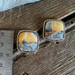 A pair of square stud earrings with silver-toned twisted rope borders and colorful landscape stone, shown on a rustic wooden surface next to a metal ruler for scale.