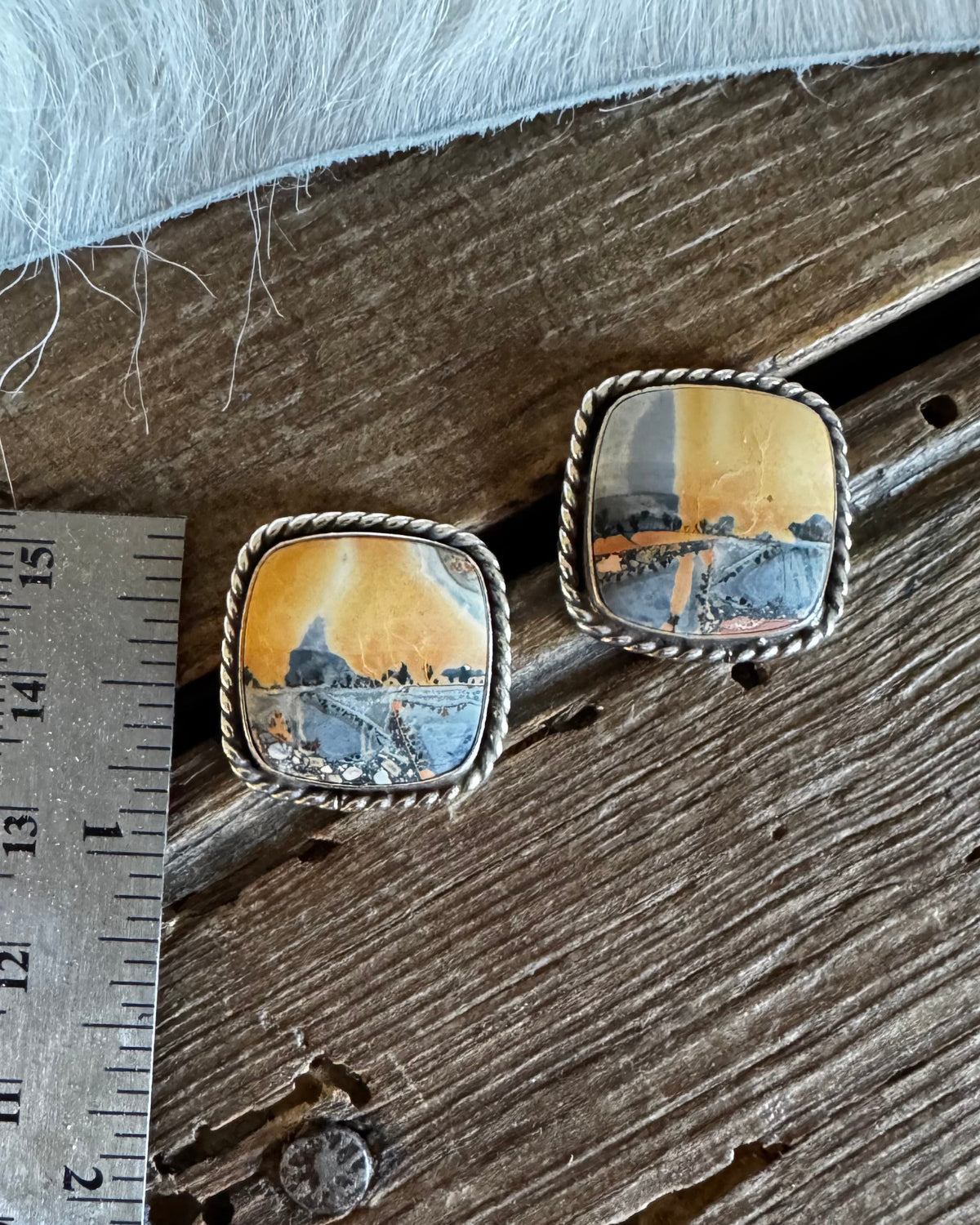 A pair of square stud earrings with silver-toned twisted rope borders and colorful landscape stone, shown on a rustic wooden surface next to a metal ruler for scale.