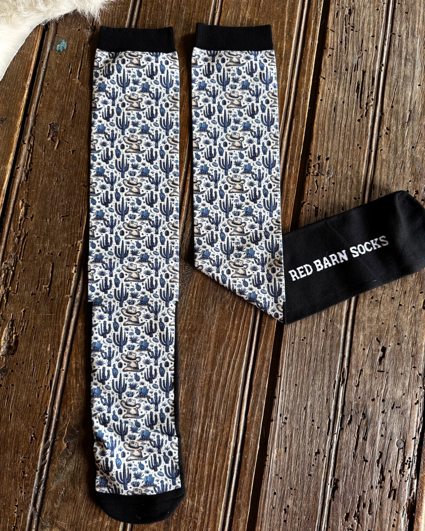 A pair of patterned crew socks laid flat on a rustic wooden surface, with one sock folded to show the black cuff and white text reading 'RED BARN SOCKS'. The socks have a blue, white, and gray floral/cactus-like pattern with black toe, heel, and cuff areas.