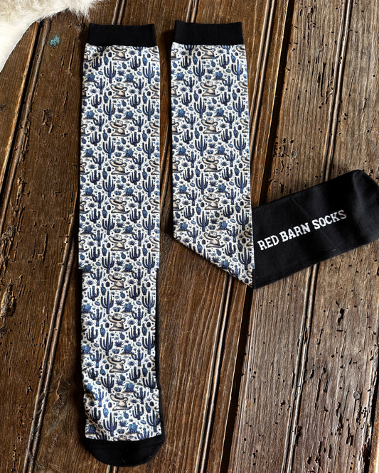 A pair of patterned crew socks laid flat on a rustic wooden surface, with one sock folded to show the black cuff and white text reading 'RED BARN SOCKS'. The socks have a blue, white, and gray floral/cactus-like pattern with black toe, heel, and cuff areas.