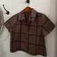 A short-sleeve button-up shirt in brown with a black grid pattern, hanging on a wooden hanger from a dark metal hook on a white wall.