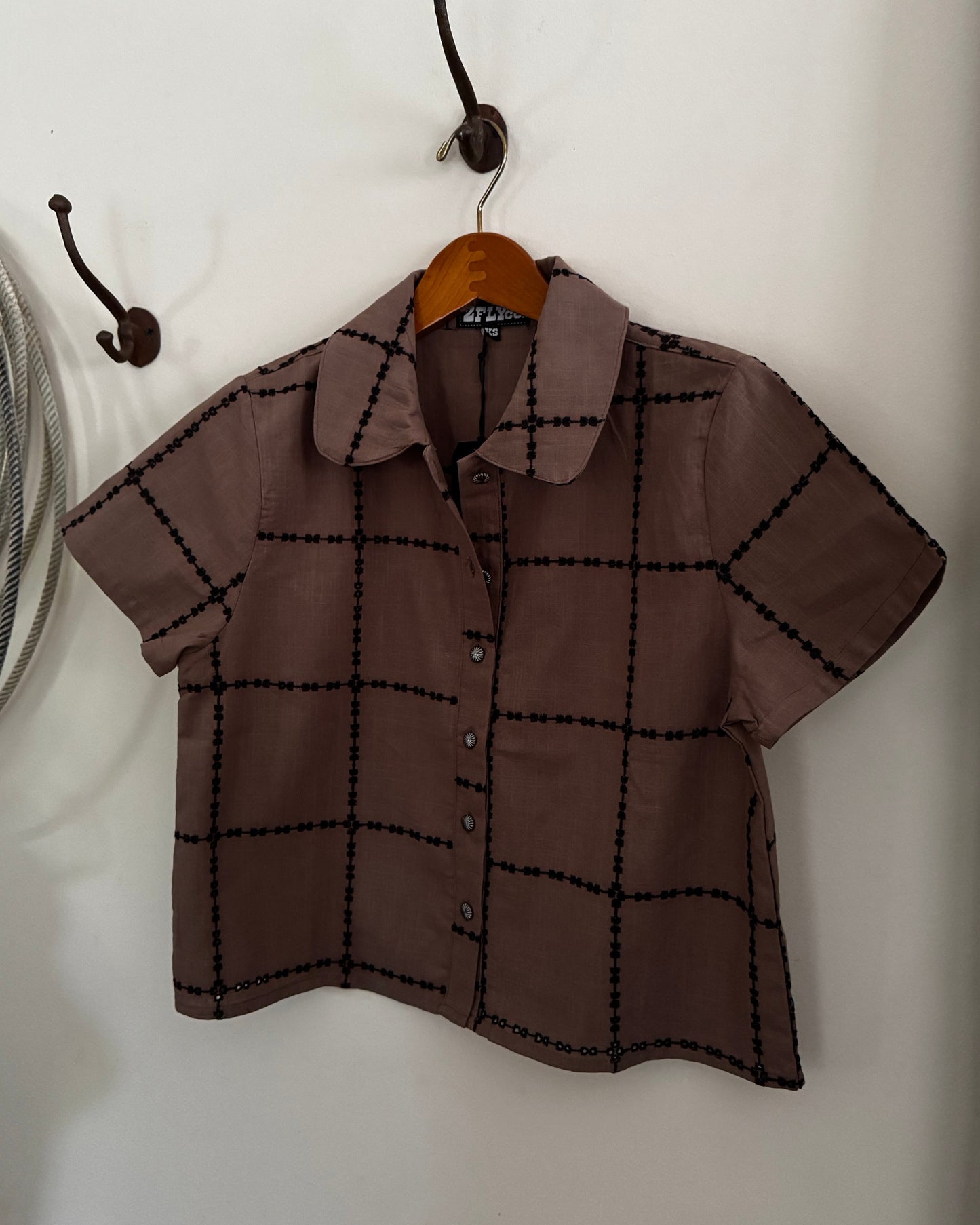 A short-sleeve button-up shirt in brown with a black grid pattern, hanging on a wooden hanger from a dark metal hook on a white wall.