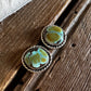 A pair of silver-toned statement earrings with oval turquoise-blue and olive-green marbled stone centers, resting on a rustic dark wood surface next to a white faux-fur trim.