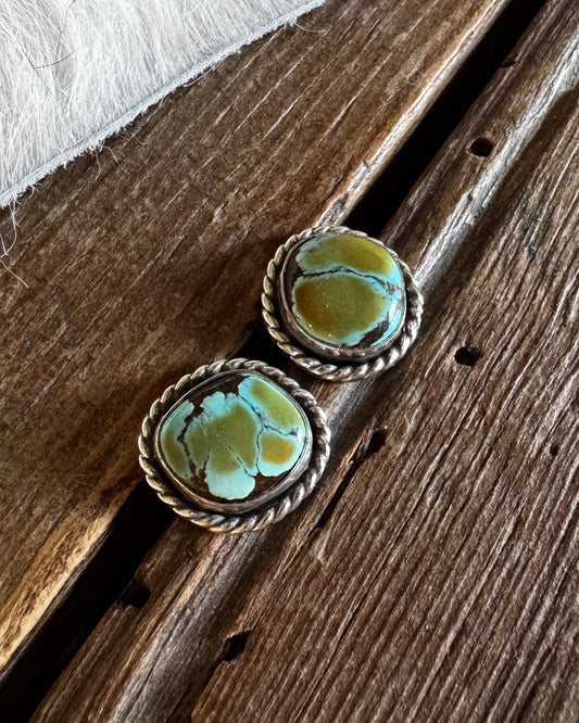 A pair of silver-toned statement earrings with oval turquoise-blue and olive-green marbled stone centers, resting on a rustic dark wood surface next to a white faux-fur trim.
