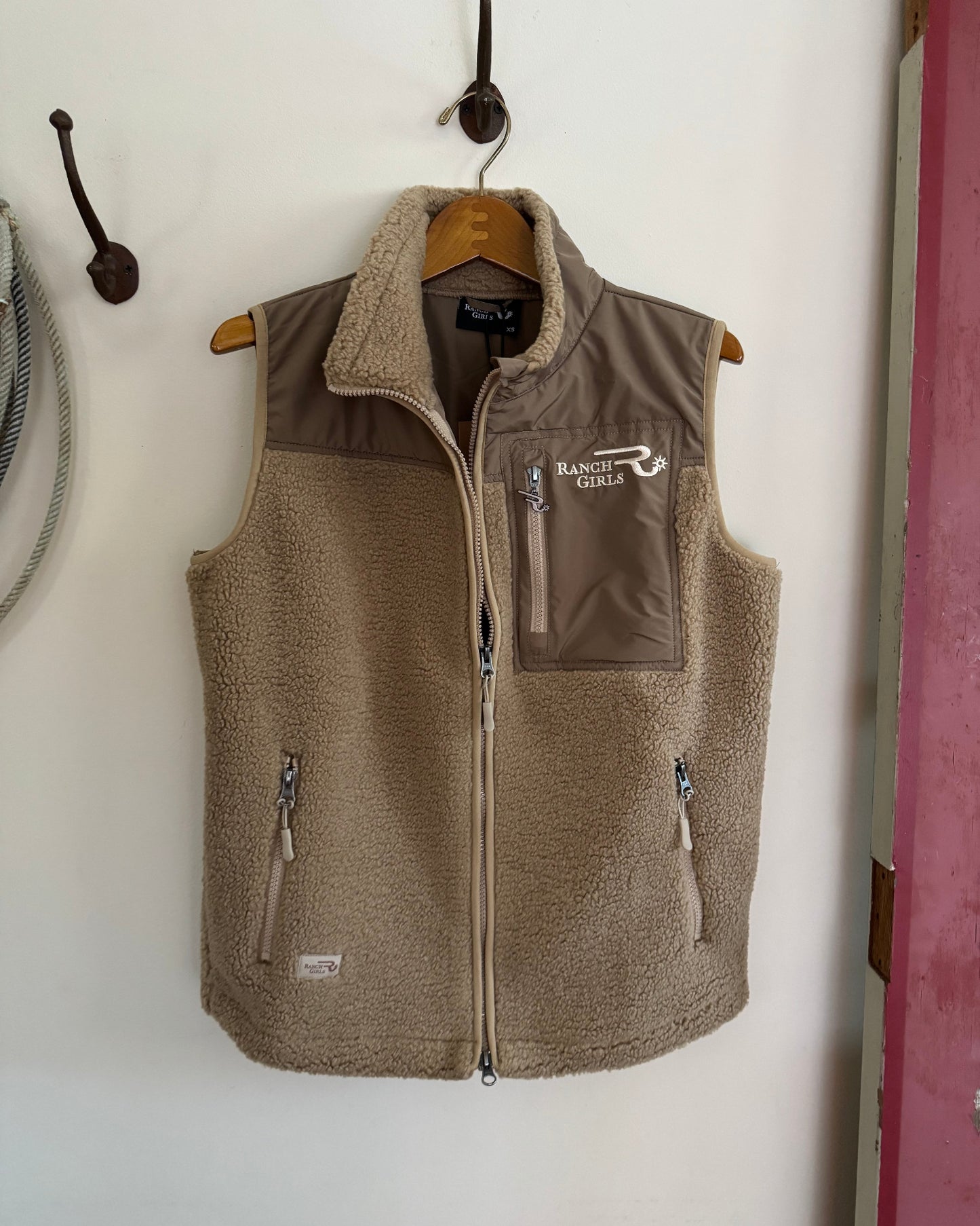 Brown vest with 'Ranch Girls' logo hanging on a hook against a white wall.
