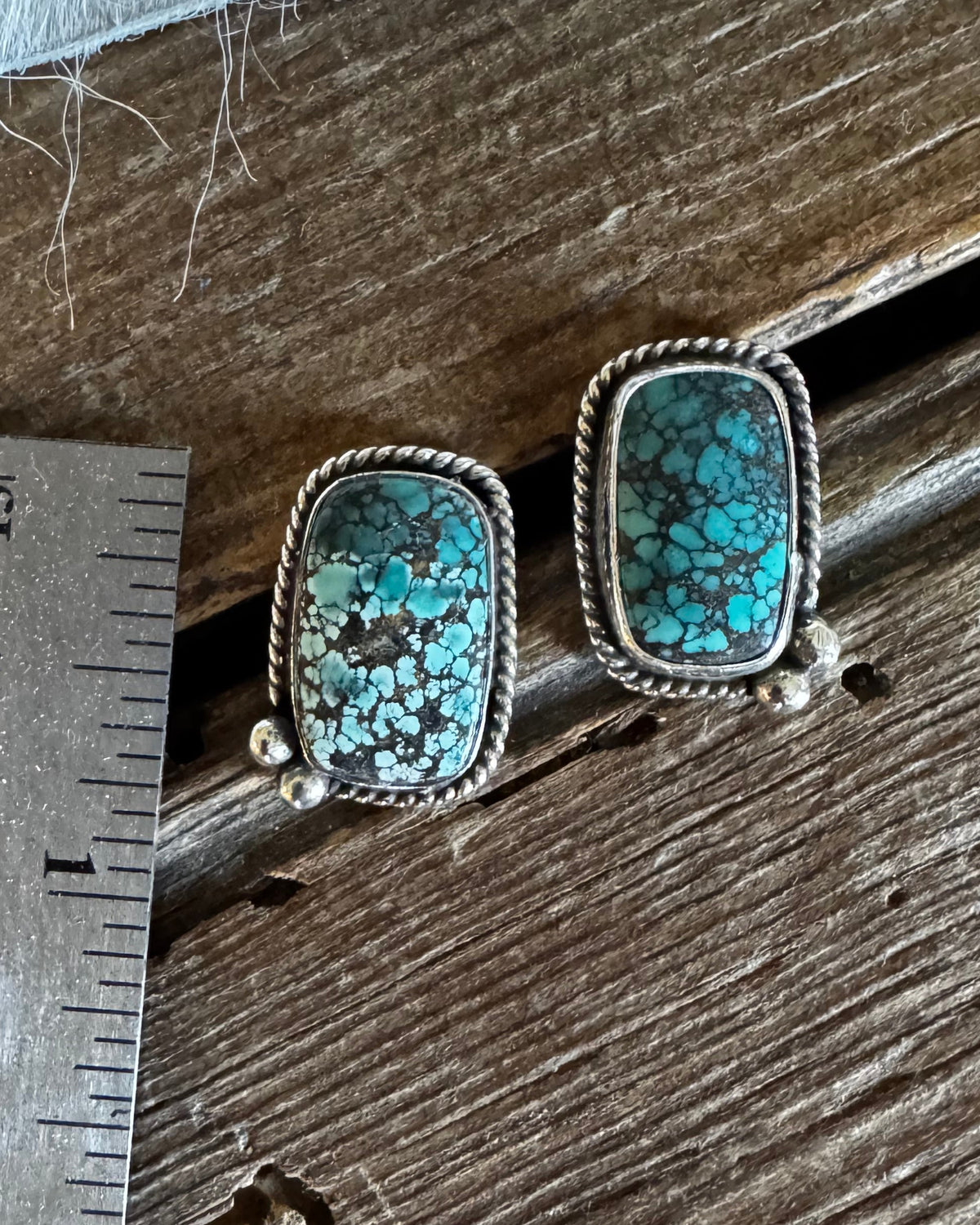 A pair of rectangular turquoise stud earrings with silver bezel settings, displayed on a rustic wooden surface next to a metal ruler for scale.
