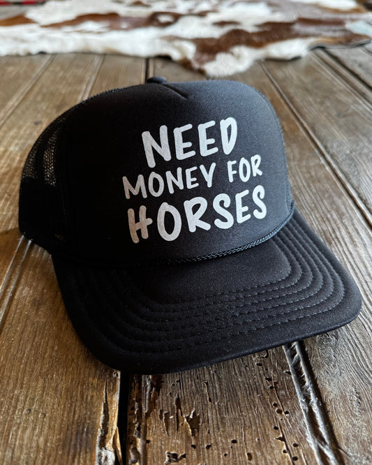 Black cap with 'Need Money for Horses' text on a wooden surface